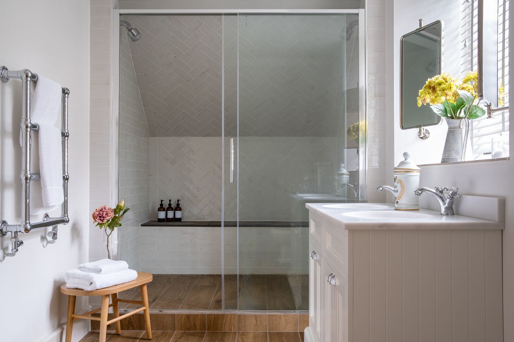 A modern bathroom with a glass shower enclosure, a wooden stool with towels, a sink with a mirror, and toiletries on the counter.