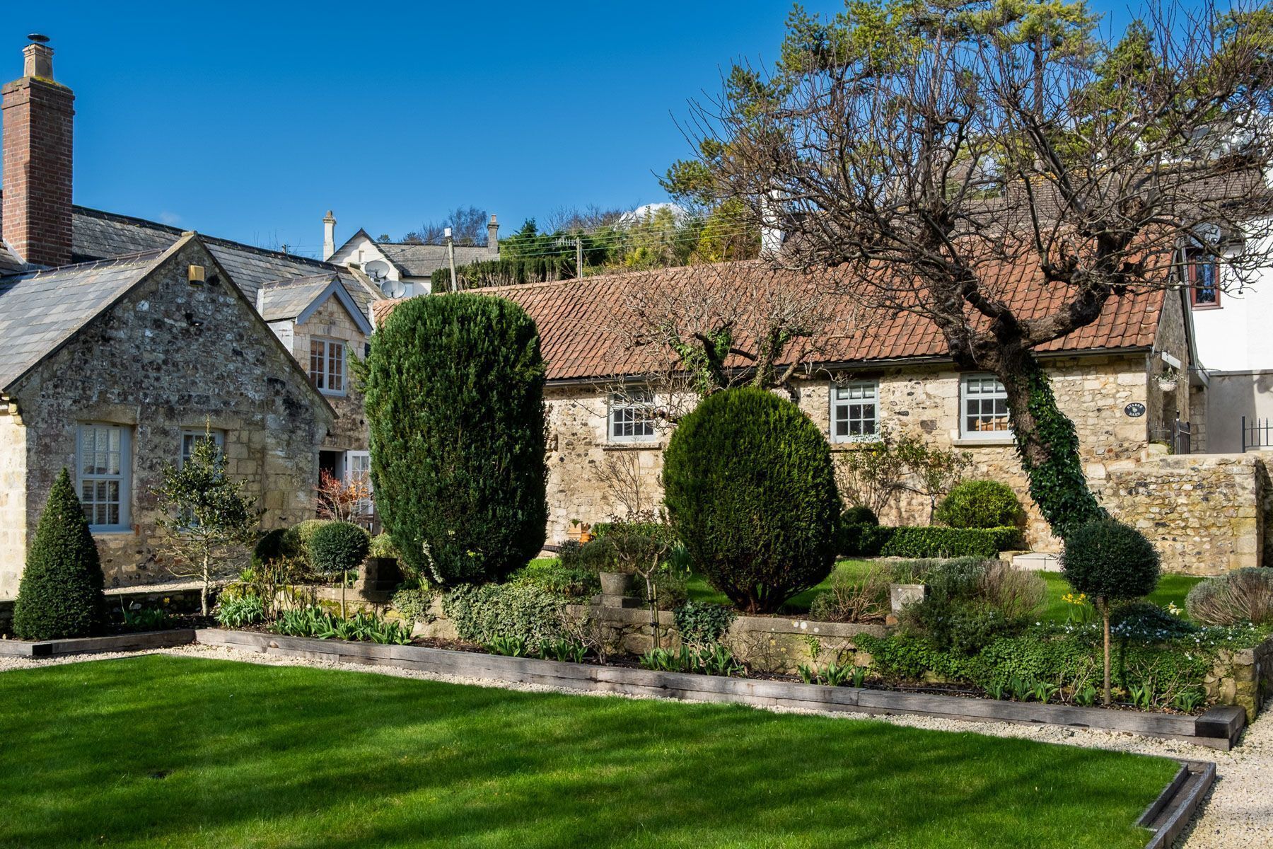 A stone house with a tiled roof, surrounded by well-maintained gardens and a clear blue sky.