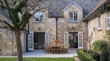 A stone building with a tiled patio, a dining table with chairs, and a tree in the foreground.