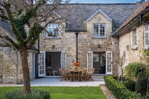 A stone building with a tiled patio, a dining table with chairs, and a tree in the foreground.