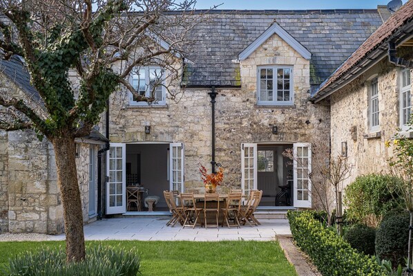 Edificio de piedra con un patio empedrado, una mesa de comedor con sillas y un árbol en primer plano.