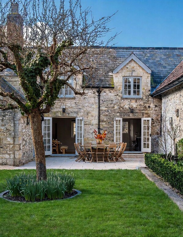 A stone building with a large tree in the foreground, a patio with a table and chairs, and a well-maintained lawn.
