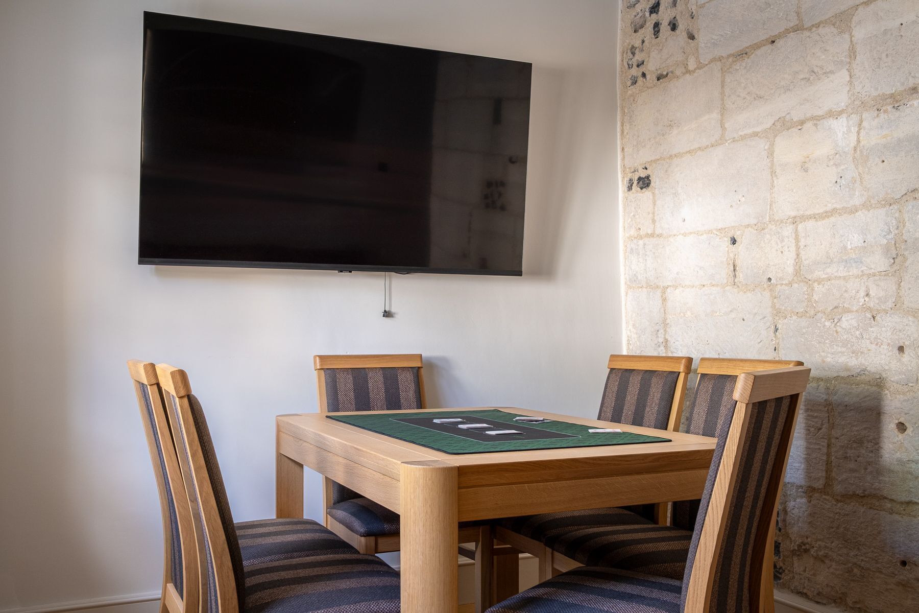 A dining area with a wooden table and chairs, a flat-screen TV mounted on the wall, and a stone wall.