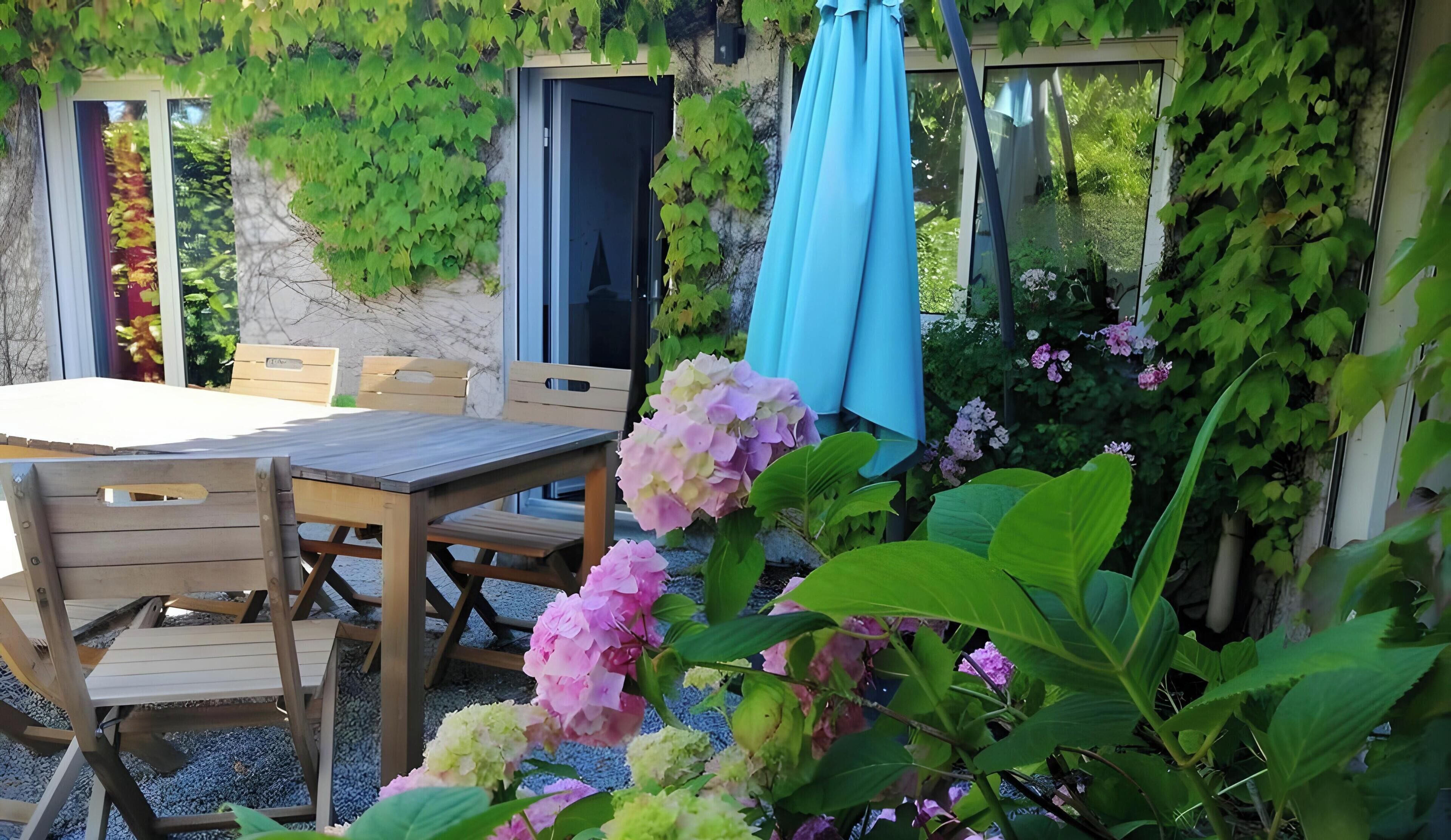 An outdoor dining area with a table and chairs, surrounded by greenery and flowering plants.