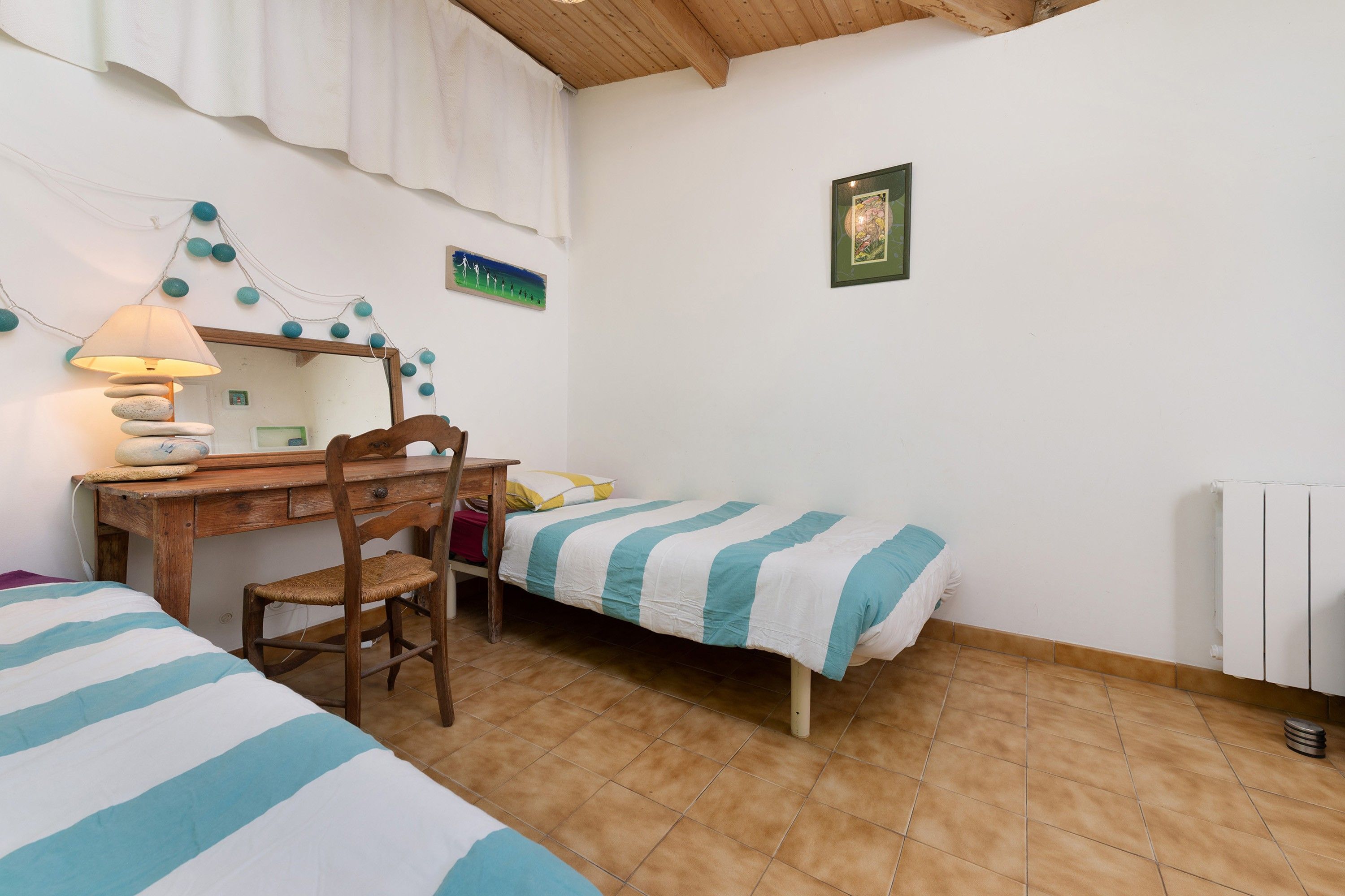 Une pièce avec un bureau en bois, une chaise, deux lits avec du linge de lit à rayures, une lampe et un tableau au mur.