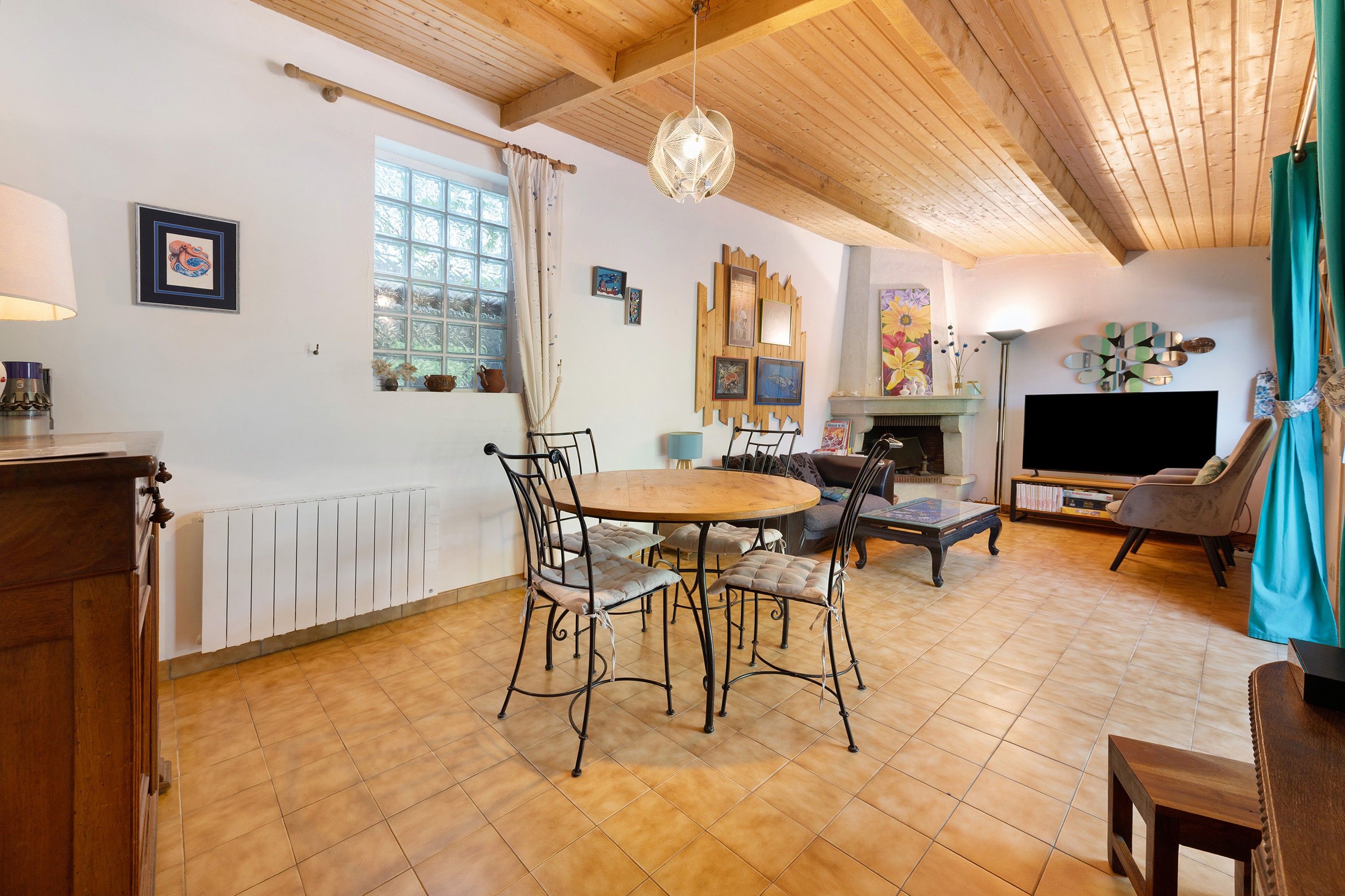 Uma sala de estar acolhedora com teto de madeira, lareira, área de jantar com mesa redonda e cadeiras, e uma televisão.