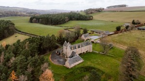 A historic stone building with a central courtyard, surrounded by rolling green fields and dense forests.