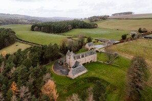A historic stone building with a central courtyard, surrounded by rolling green fields and dense forests.