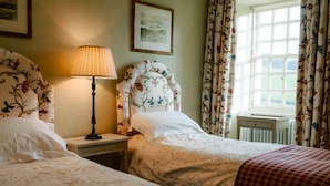 A bedroom with two beds, a bedside table with a lamp, floral curtains, and a window with a view.