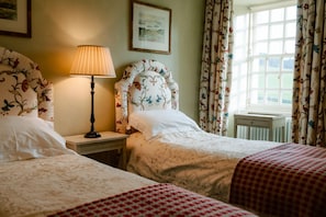 A bedroom with two beds, a bedside table with a lamp, floral curtains, and a window with a view.