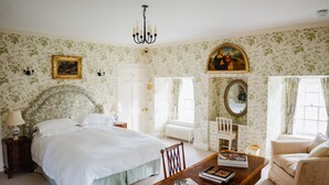 Une chambre avec un grand lit, une table de chevet en bois, une commode, une chaise, une table avec des livres, un lustre, une fenêtre avec des rideaux, un radiateur et un tableau au mur.