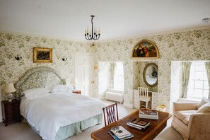 Una camera da letto con un letto grande, un comodino in legno, una cassettiera, una sedia, un tavolo con libri, un lampadario, una finestra con tende, un termosifone e un quadro appeso al muro.