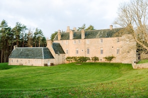 A large stone building with multiple chimneys, a well-maintained lawn, and a forested background. - Finnie Castle - Nine Bedroom Castle, Sleeps 18 (Jedburgh)