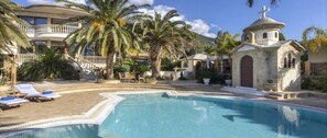 A pool area with lounge chairs, palm trees, and a small church-like structure. - Villa Verity - Nine Bedroom Villa, Sleeps 21 (Porto Germeno)