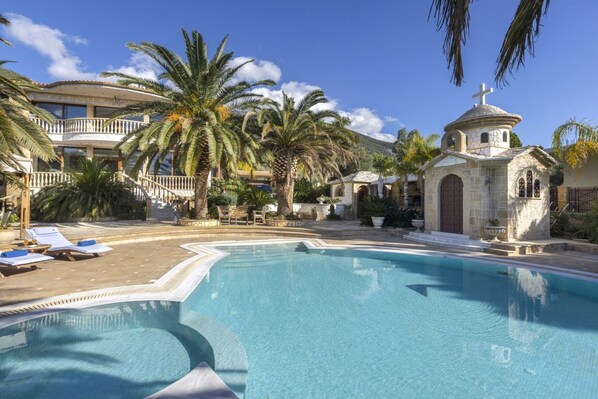 A swimming pool with lounge chairs, palm trees, and a small church with a cross on top. - Villa Verity - Nine Bedroom Villa, Sleeps 21 (Porto Germeno)