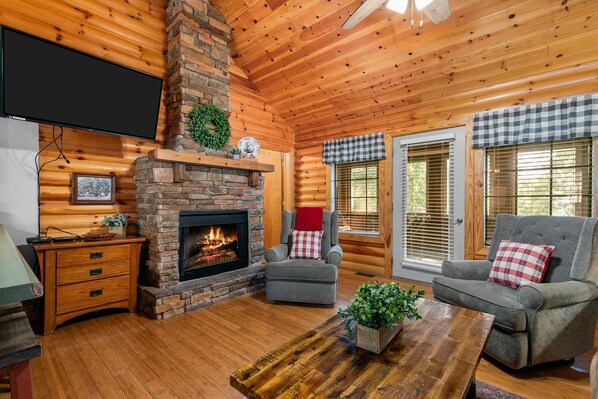 A cozy cabin interior with a stone fireplace, wooden walls, a television, and two gray sofas with plaid cushions.