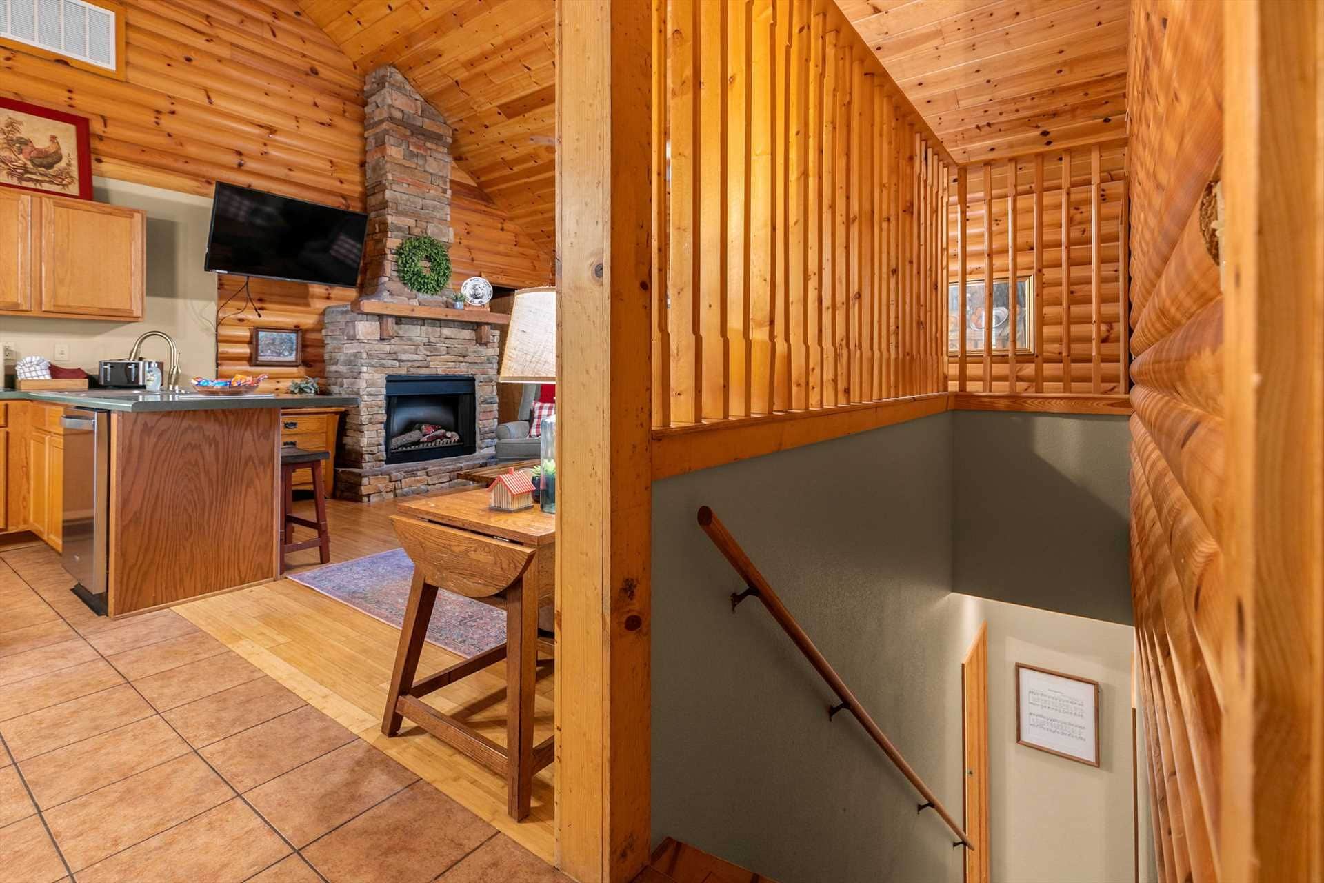 A cozy cabin interior with a stone fireplace, wooden walls, a kitchen area, and a large window.