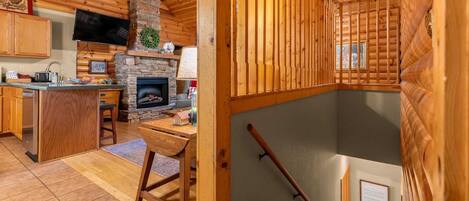 Interior acogedor de una cabaña con chimenea de piedra, paredes de madera, zona de cocina y un ventanal grande.
