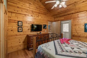Quarto de cabana de madeira com cama, cômoda, TV e ventilador de teto.