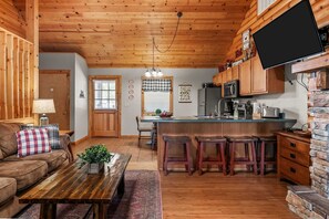 A cozy cabin interior with a wooden ceiling, a dining area with a wooden table and bar stools, a kitchen with a refrigerator and microwave, and a television mounted on a stone wall. - Quiet Cabin1 mile to SDC - 4 Private Bed/Bath Masters, 2 Living Areas, 2 Pools! | 35 (Branson West)