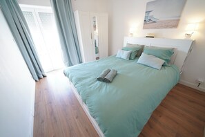 A neatly made bed with a light-colored bedspread and pillows, a headboard with a light fixture, and a view of the beach through a window. - Cascais apartment with 2 bedrooms, 70 m², and a sunny terrace! (Cascais)