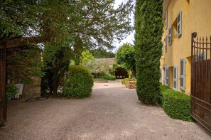 Uma entrada de cascalho leva a um edifício com fachada amarela e persianas, cercado por vegetação e muros de pedra.