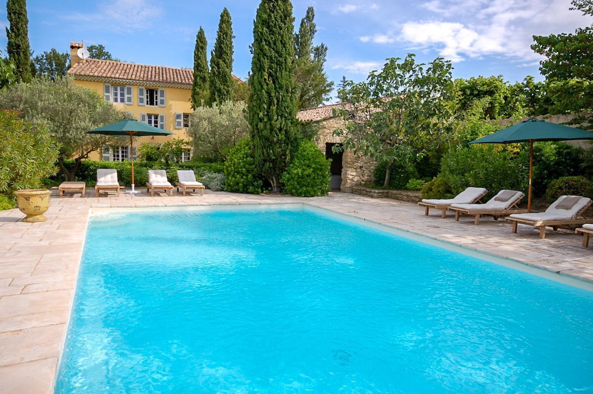 A rectangular pool with lounge chairs and umbrellas, surrounded by greenery and a building with a tiled roof.