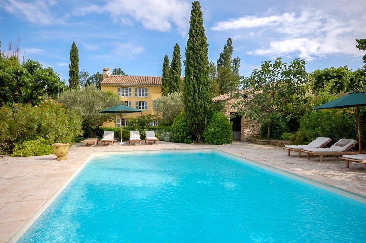 A swimming pool with lounge chairs and umbrellas, surrounded by greenery and a building with a tiled roof.