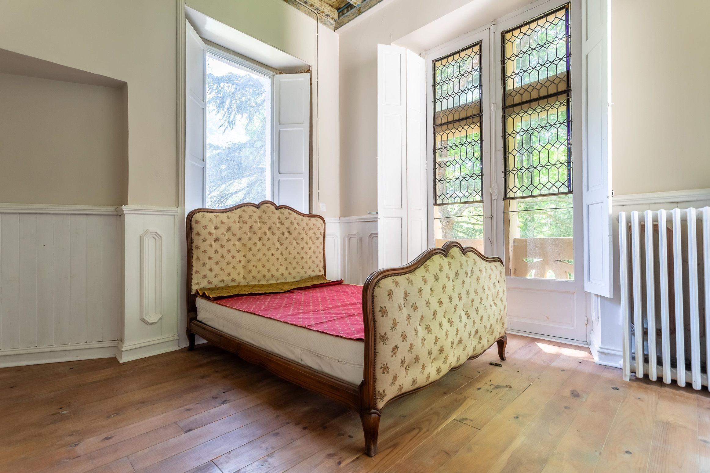 Une pièce avec un plancher en bois, un lit simple doté d’une tête de lit à motifs floraux et une fenêtre avec une grille en métal.