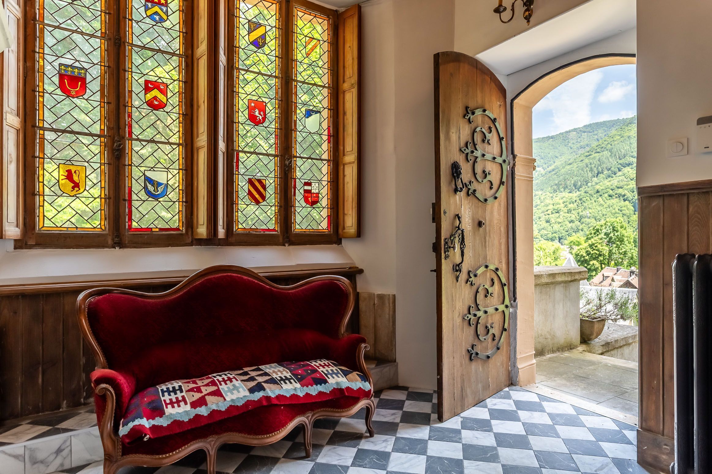 A room with a red velvet sofa, a checkered floor, and a large window with stained glass featuring various coats of arms.