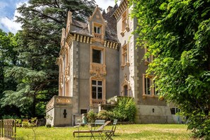 Een historisch gebouw met een opvallende toren, een balkon en een tuin met zitplaatsen.