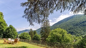 A grassy outdoor area with a metal fence, a table with chairs, and a view of a forested mountain.