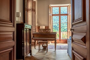 A grand piano in a spacious room with wooden paneling and a large window offering a view of a forest. - Chateau Des Pyrenees (Bagnères-de-Luchon)