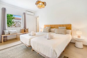 Habitación de hotel con dos camas, un banco, una ventana que ofrece vista a un muro de piedra y follaje, y una lámpara de araña moderna.