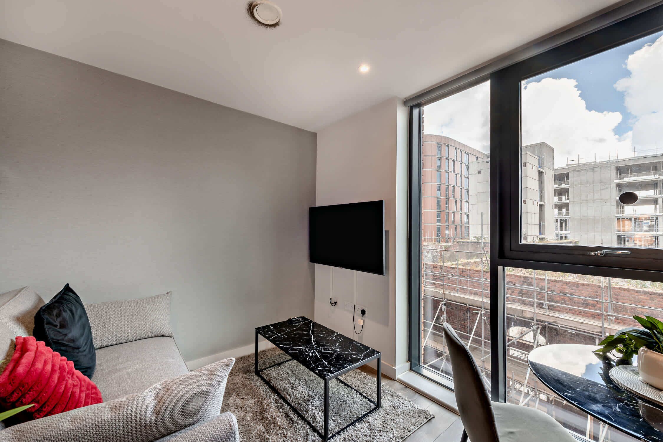 A modern living room with a large window, a flat-screen TV mounted on the wall, a small black table, and a sofa with decorative pillows.
