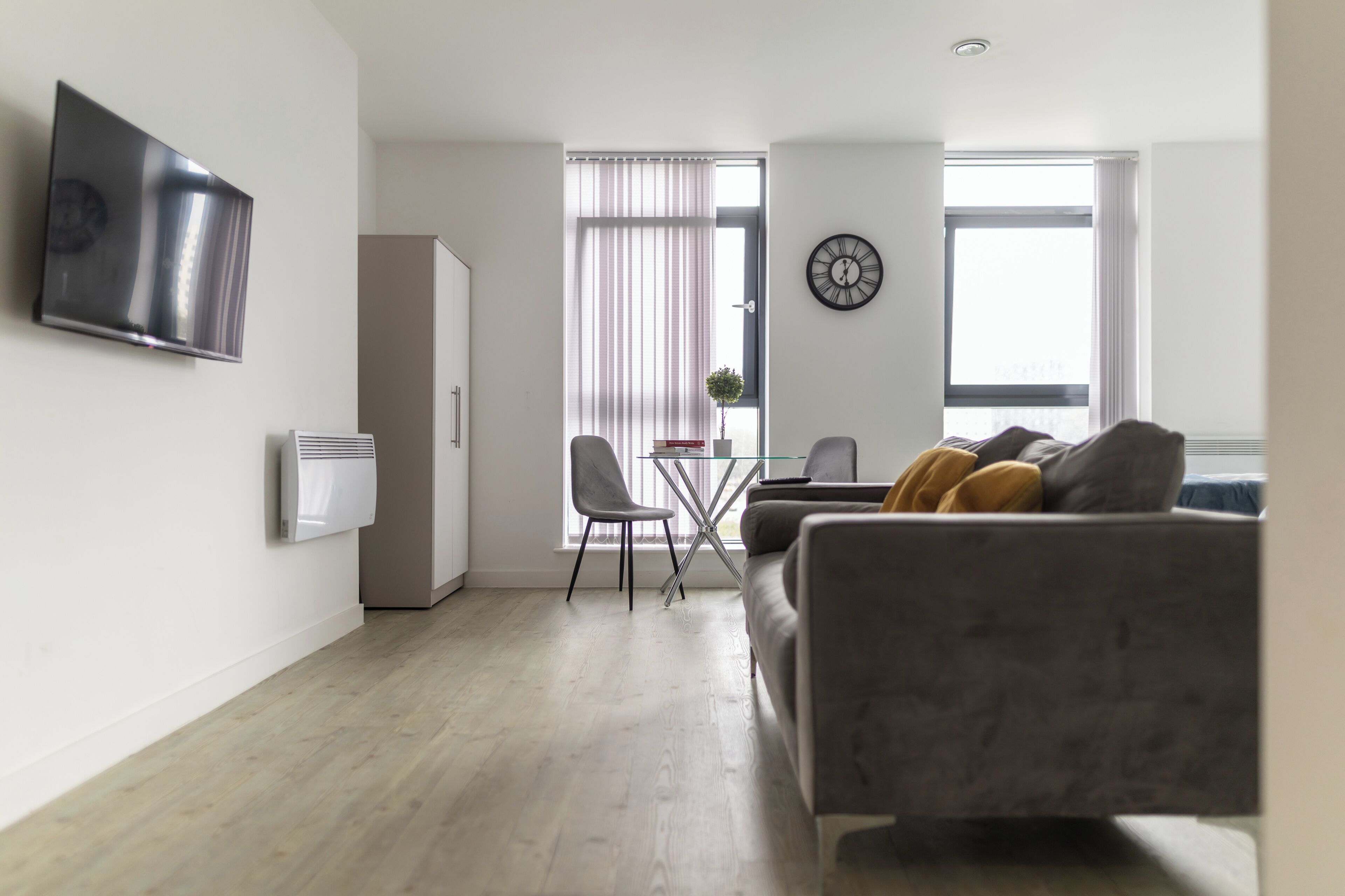 A modern living room with a grey sofa, a flat-screen TV mounted on the wall, and a dining area with a small table and chairs.
