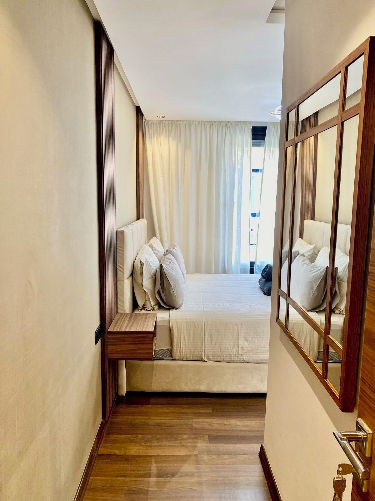 A narrow hallway leading to a bedroom with a bed, bedside table, and a mirror.