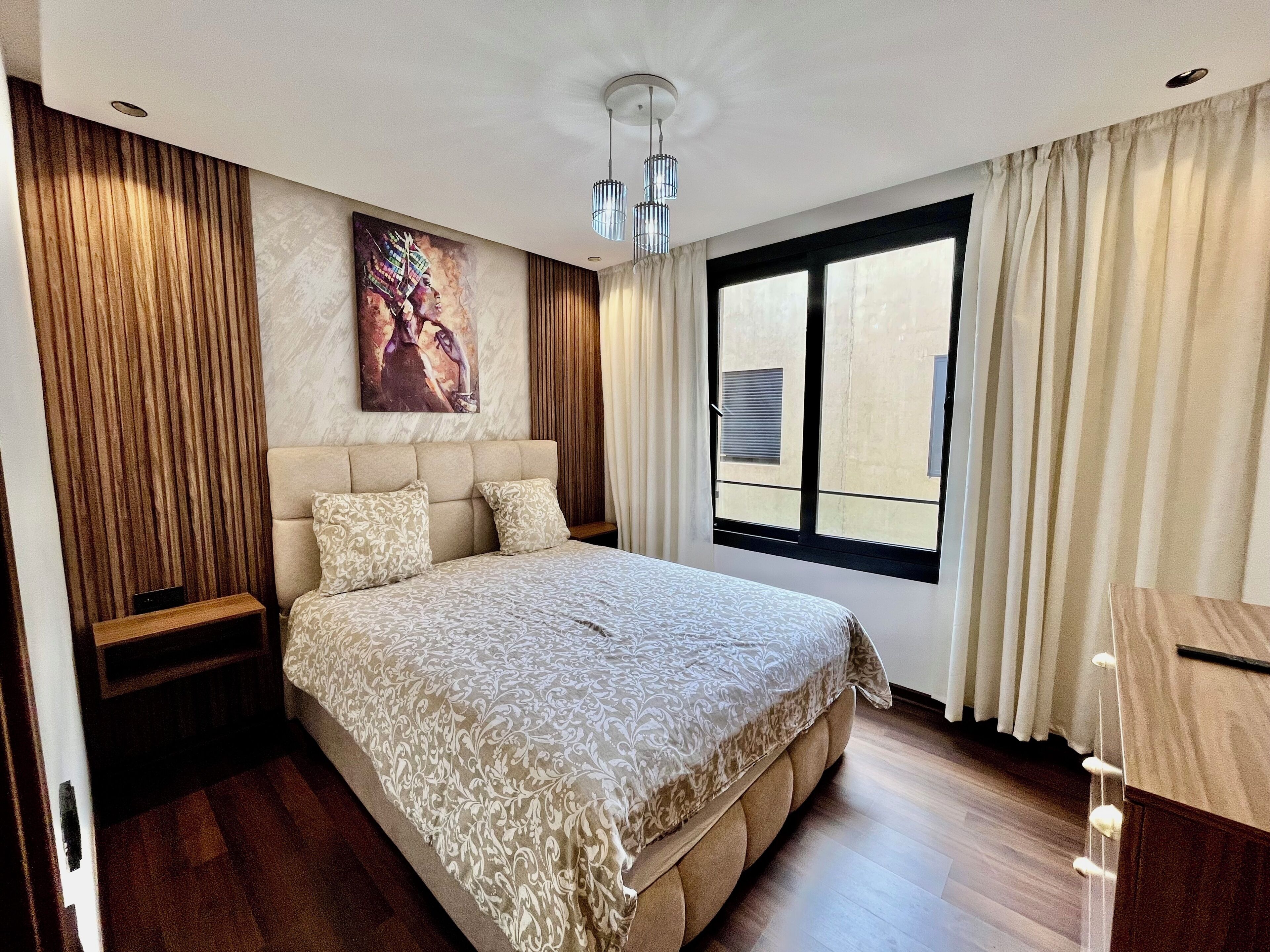A modern bedroom with a large bed, a headboard, a window with curtains, and a painting on the wall.
