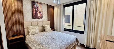 A modern bedroom with a large bed, a headboard, a window with curtains, and a painting on the wall.