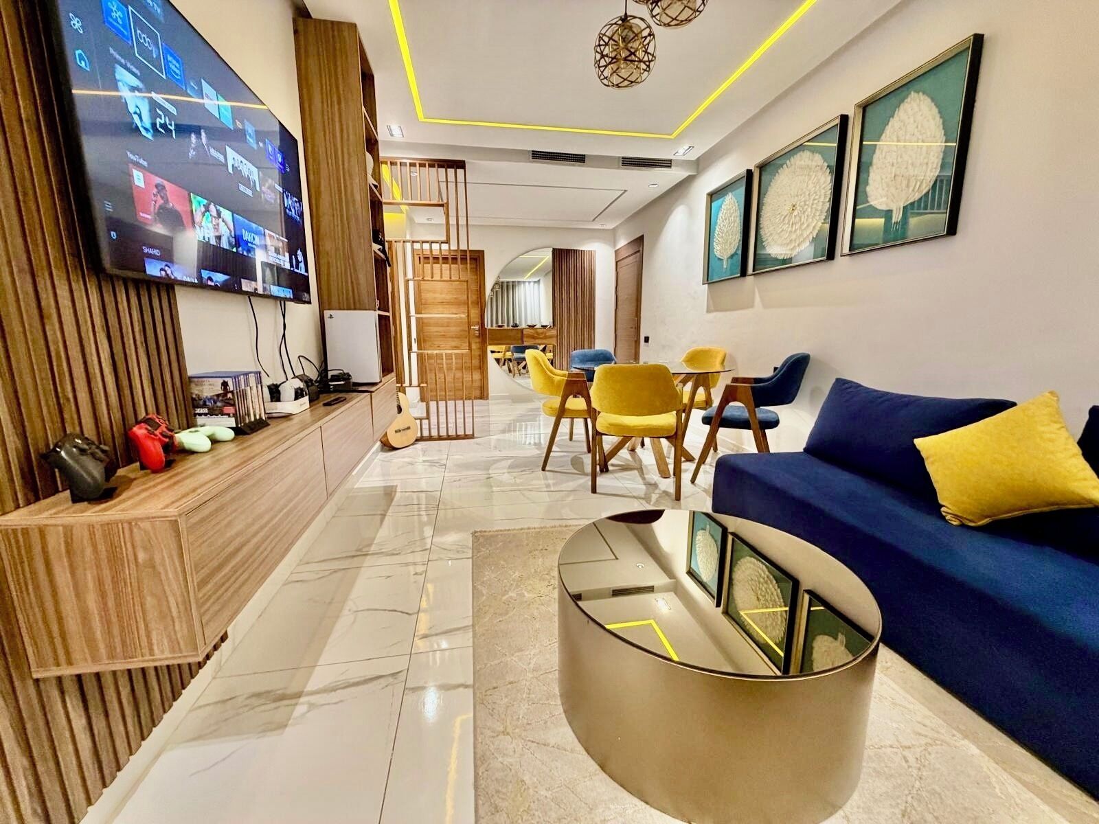 A modern living room with a flat-screen TV, a blue sofa, yellow chairs, and a round coffee table.