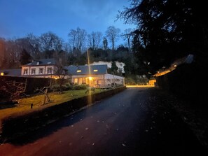 A two-story building with a stone facade, a paved driveway, and a small stream running alongside. - La Cour de Flaubert – Large vacation rental for 25 people near Rouen (Morville-le-Héron)