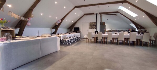 An indoor venue with a high ceiling and exposed wooden beams, featuring a long table set for a meal and a separate dining area with chairs. - La Cour de Flaubert – Large vacation rental for 25 people near Rouen (Morville-le-Héron)