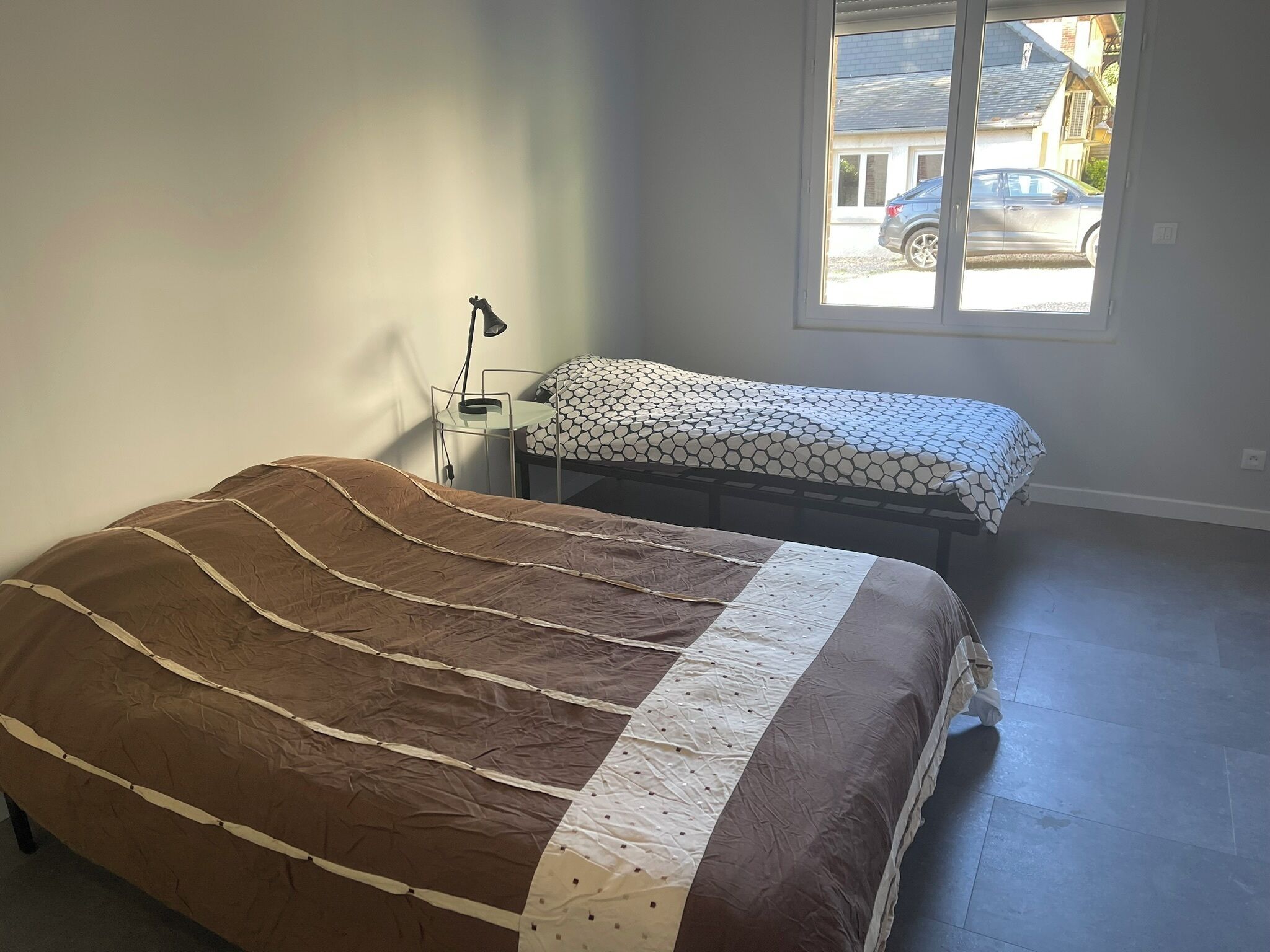 Une chambre à coucher avec un lit, une table de chevet, une lampe et une fenêtre donnant sur une voiture garée à l’extérieur.