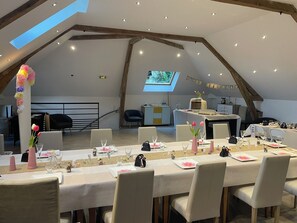 An indoor dining area with a high ceiling and exposed beams, a long table set for a meal, and a television screen in the background. - La Cour de Flaubert – Large vacation rental for 25 people near Rouen (Morville-le-Héron)