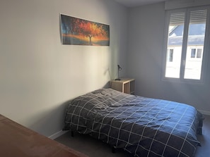 A bedroom with a bed, a nightstand, a lamp, a window, and a painting on the wall.