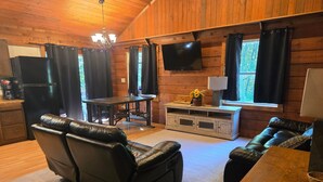 A cozy cabin interior with a wooden ceiling, a flat-screen TV mounted above a window, a dining table with chairs, and a leather sofa. - Lakefront views of Willscreek Lake 2 bedroom (Adamsville)