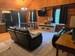 Ein gemütliches Blockhaushaus-Interieur mit Holzbalkendecke, einem flachbildfernseher über einem Fenster, einem Esstisch mit Stühlen und einem Ledersofa.