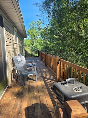 Eine Holzterrasse mit zwei Stühlen und einem Grill, umgeben von Bäumen und einem Haus.