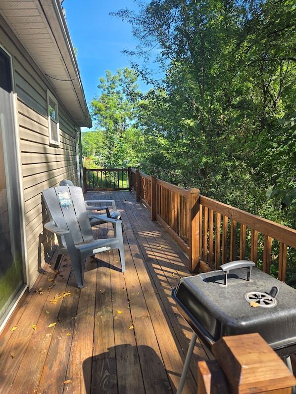 A wooden deck with two chairs and a grill, surrounded by trees and a house.