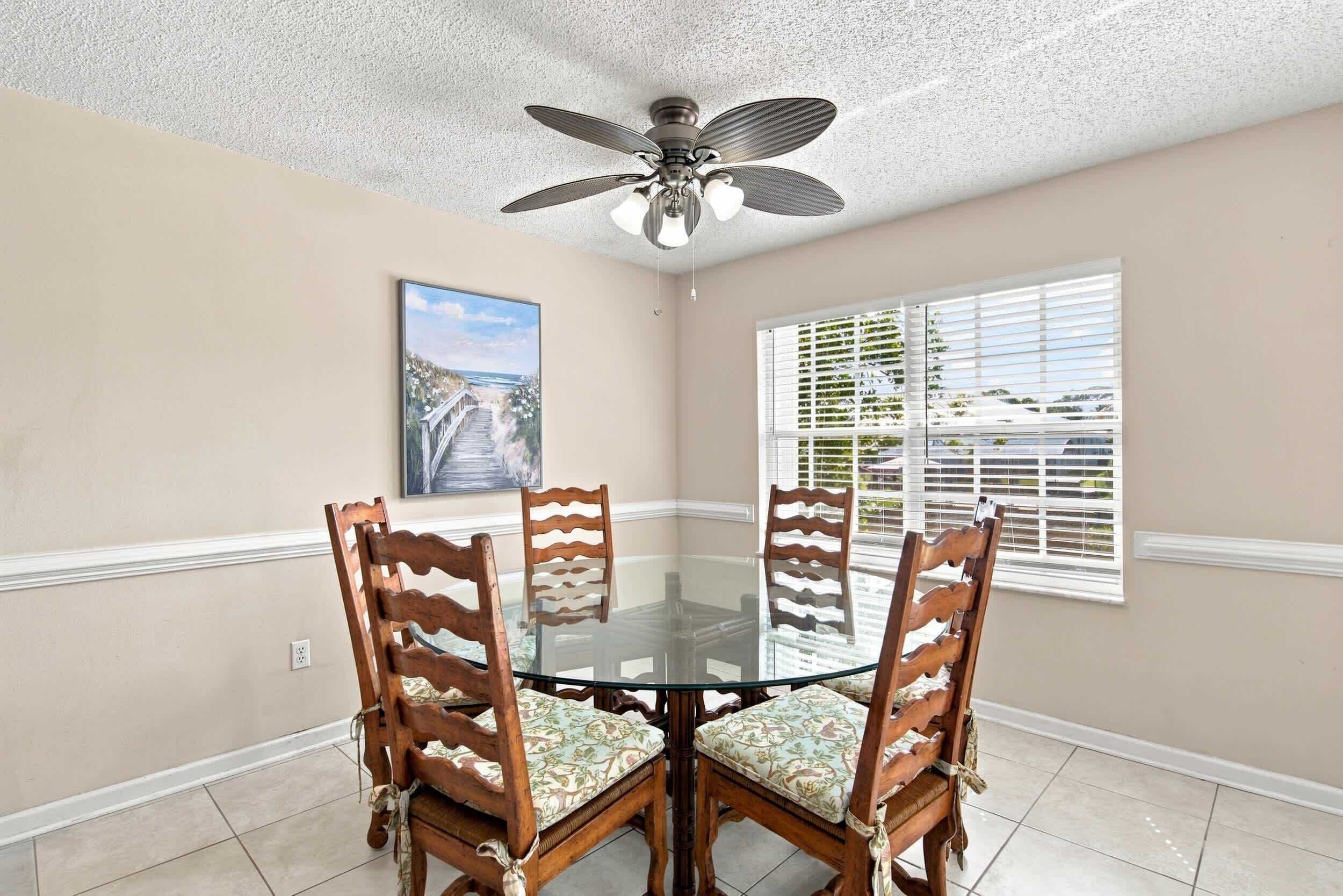 A dining area with a glass table surrounded by wooden chairs with cushioned seats, a ceiling fan, a painting on the wall, and a window with blinds.